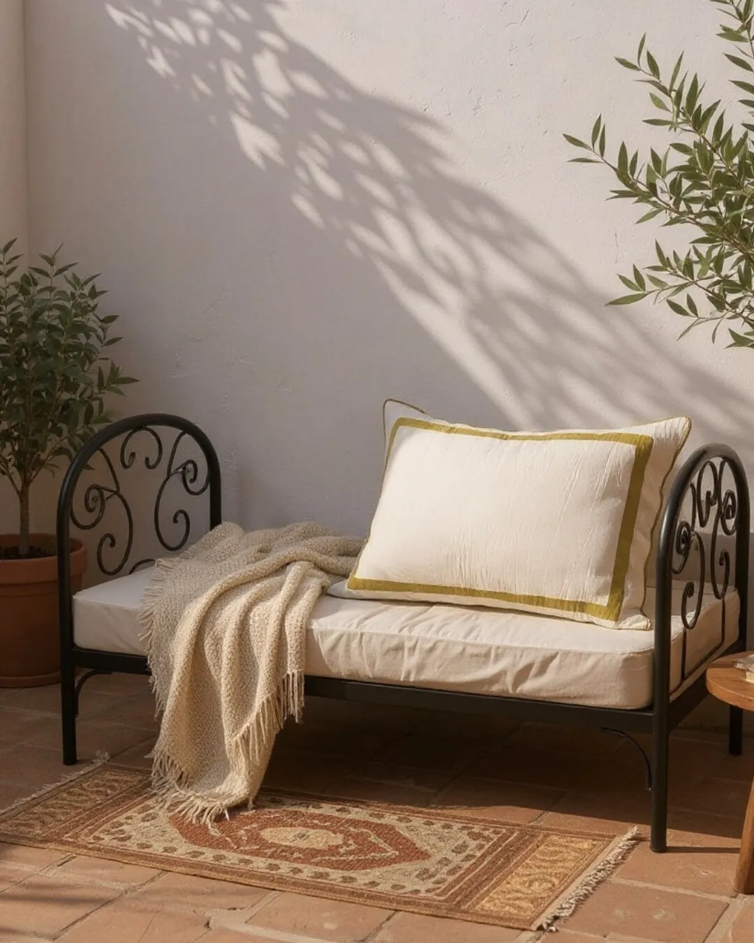 Diván de hierro negro con almohadón bordado y manta tejida en un patio soleado con plantas.
