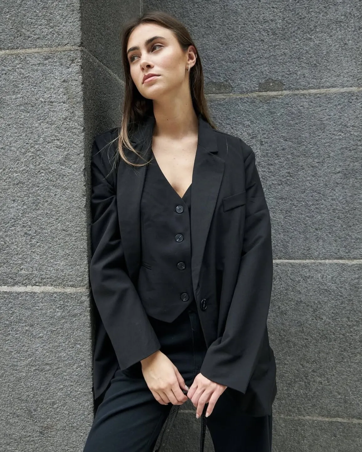 Modelo con blazer y chaleco negros apoyada contra una pared de piedra gris.